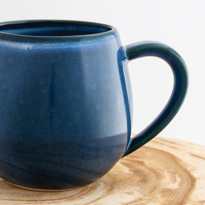 Close up of a Robert Gordon dark topaz blue canvas mug coffee cup sitting on a natural wooden round side table